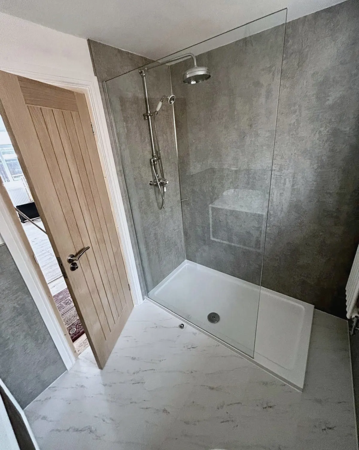 Walk-in shower with a frameless glass screen and a rainfall shower head, beside an oak door